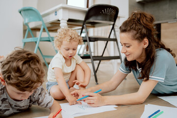 The family spends their free time together. A mother with her school-age son and preschool-age daughter draw together while sitting on the wooden floor in the living room at home.