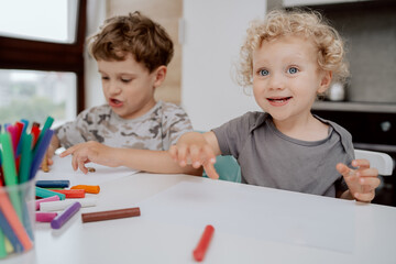 The happy siblings are sitting at the kitchen table playing with plasticine. A school-aged boy and a preschool-aged girl spend their free time together.