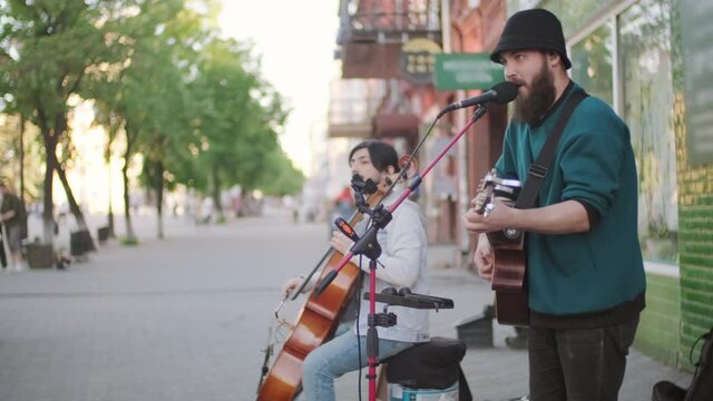 Side view shot of male street musicians performing song outdoors. One man playing cello while the other playing acoustic guitar and singing into microphone