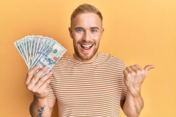 Young caucasian man holding dollars pointing thumb up to the side smiling happy with open mouth