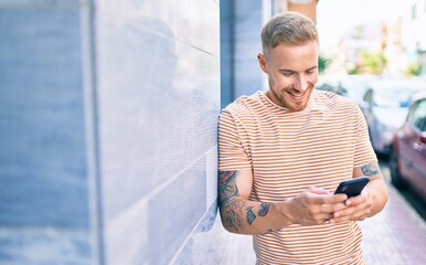 Young irish man smiling happy using smartphone leaning on the wall at street of city.