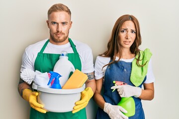 Young couple of girlfriend and boyfriend wearing apron holding products and cleaning spray clueless and confused expression. doubt concept.