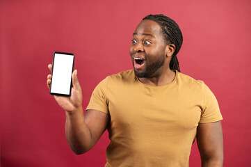 Surprised by the possibilities of new mobile app African-American man staring at smartphone with empty screen, astonished dark skinned guy demonstrates phone with blank display, isolated on red