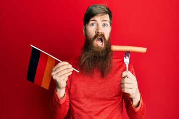 Redhead man with long beard holding fork with pork sausage and germany flag in shock face, looking...