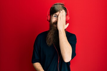 Redhead man with long beard listening to music using headphones covering one eye with hand, confident smile on face and surprise emotion.