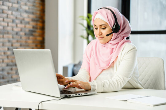 Young Muslim Arab mixed-race female customer service representative in hijab and headset sitting at the desk in office, typing email on laptop, searching or browsing the internet, replying to client