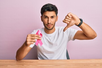 Young handsome man holding pink cancer ribbon with angry face, negative sign showing dislike with...