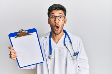 Young handsome man wearing doctor stethoscope holding clipboard scared and amazed with open mouth...