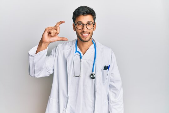 Young handsome man wearing doctor uniform and stethoscope smiling and confident gesturing with hand doing small size sign with fingers looking and the camera. measure concept.