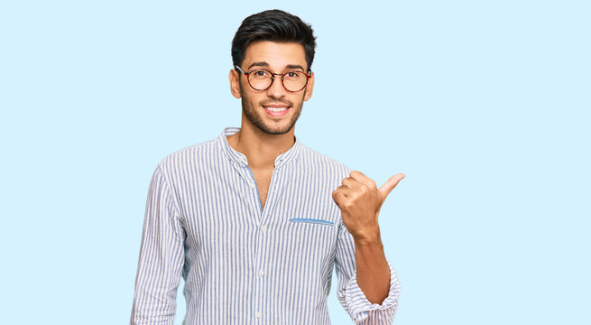 Young handsome man wearing casual clothes and glasses smiling with happy face looking and pointing to the side with thumb up.