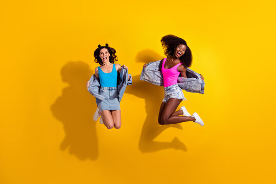 Photo of two crazy active ladies jump open mouth wear jeans shorts skirt jacket top shoes isolated yellow color background
