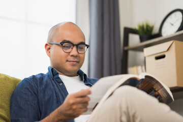 Young asian man sitting and reading book on sofa in living room.