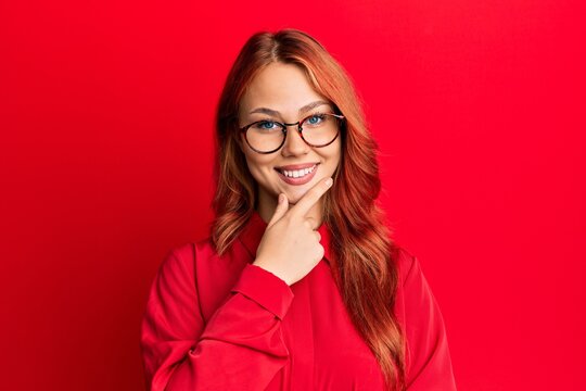 Young beautiful redhead woman wearing casual clothes and glasses over red background looking confident at the camera smiling with crossed arms and hand raised on chin. thinking positive.