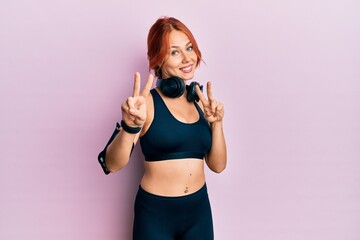 Young beautiful redhead woman wearing gym clothes and using headphones smiling looking to the camera showing fingers doing victory sign. number two.