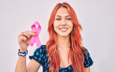 Young redhead girl smiling happy holding breast cancer pink ribbon walking at the city.