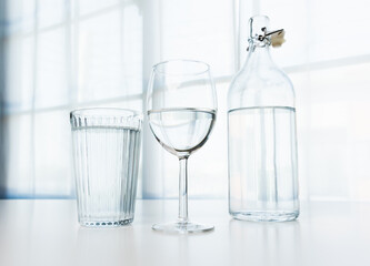 A glass of water and a clear glass water bottle are placed on the table in the house on a white background.