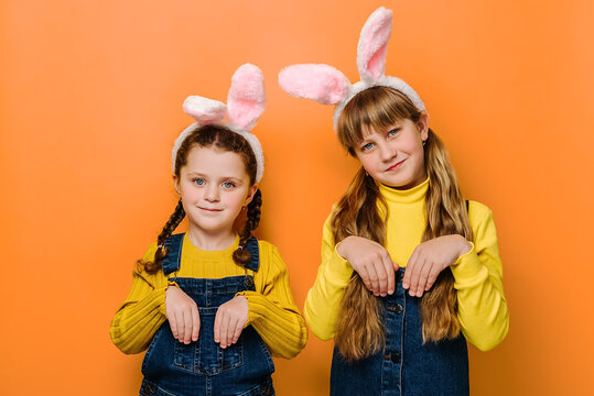 Charming lovely little girls with friendly expression, beautiful sisters keeps hands as if rabbit, wears pink bunny fluffy ears, isolated on orange studio background. Happy Easter holiday concept