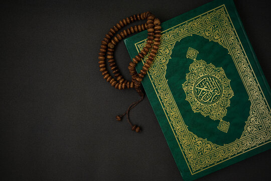 Holy Al Quran with written arabic calligraphy meaning of Al Quran and rosary beads or tasbih on black background with a copy space. Flat lay.