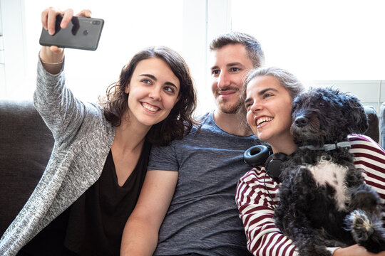 A group of happy young friends hugging their affectionate dog and having their picture taken with the phone in a bright room. Selfie of young family. Selfie of group of friends