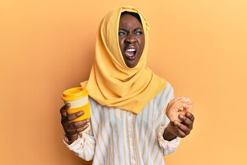Beautiful african young woman wearing islamic hijab drinking a coffee using smartphone angry and mad screaming frustrated and furious, shouting with anger. rage and aggressive concept.