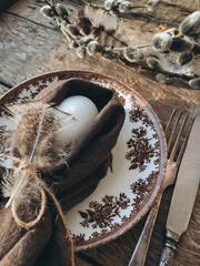 Easter egg in napkin, feathers, vintage plate, cutlery on aged wood. Rustic Easter table setting