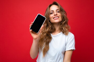 Photo of beautiful smiling young blonde woman good looking wearing white t-shirt standing isolated on red background with copy space holding phone showing smartphone in hand with empty screen display