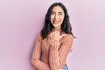 Hispanic teenager girl with dental braces wearing casual clothes smiling with happy face looking and pointing to the side with thumb up.