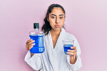 Hispanic teenager girl with dental braces holding mouthwash puffing cheeks with funny face. mouth inflated with air, catching air.