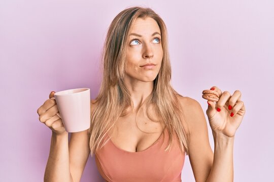 Beautiful caucasian woman drinking coffee and eating french pastry macaron smiling looking to the side and staring away thinking.