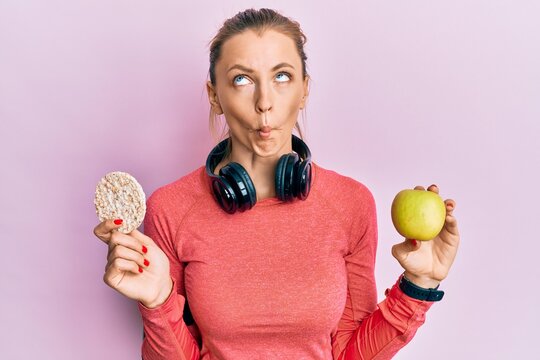 Beautiful caucasian sports woman holding green apple and rice crackers making fish face with mouth and squinting eyes, crazy and comical.