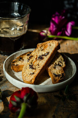 Homemade almond and chocolate biscotti with glass of coffee and  ranunculus flowers on wooden table on dark background.