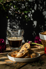 Homemade almond and chocolate biscotti with glass of coffee and  ranunculus flowers on wooden table on dark background.