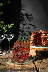 Piece of homemade chocolate and cherry cake, whole cake  on wooden stand, glasses and flowers on dark background.