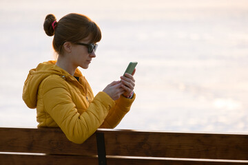 Young pretty woman sitting on a park bench browsing her sellphone outdoors in the evening.