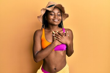 Young african american woman wearing bikini and summer hat smiling with hands on chest, eyes closed with grateful gesture on face. health concept.