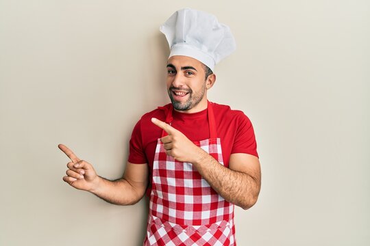 Young hispanic man wearing baker uniform smiling and looking at the camera pointing with two hands and fingers to the side.