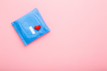 Bright red heart on blue pack of sanitary towel on light pink table background. Pastel color. Closeup. Protection in day and night time. Empty place for text. Top down view. Copy space.