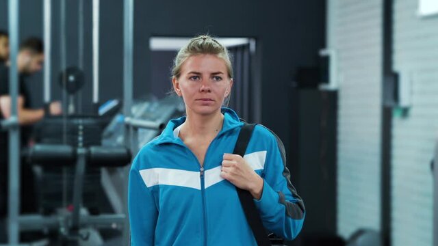 Young woman wearing blue sports jacket walking in gym with bag on shoulder. Tracking shot athletic female before training. Concept of healthy lifestyle