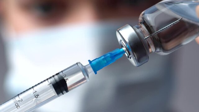 Doctor in a white medical mask and blue gloves holds in his hands a syringe and a vaccine bottle at the hospital. Physician dials the medicine into the syringe