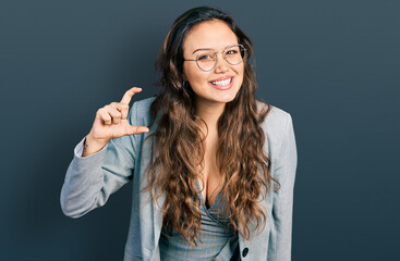 Young hispanic girl wearing business clothes and glasses smiling and confident gesturing with hand doing small size sign with fingers looking and the camera. measure concept.