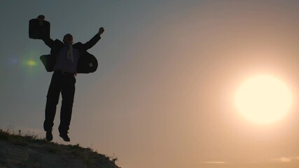 A business man at sunset, an entrepreneur jumps on the top of a mountain at sunset, rejoicing by raising his hand up a symbol of success, a successful deal, career growth. The concept of teamwork.
