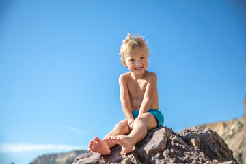 A blond little boy in swimming shorts sits on a large rock against the blue sky.