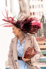 A woman with pink curly hair blowing in the wind. Standing on the stairs in a light raincoat and...