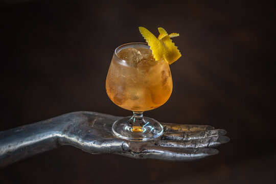 cocktail ghiacciato all'arancia in bicchiere da whisky, decorato con buccia d'arancia, poggiato su scultura a forma di mano di ferro 