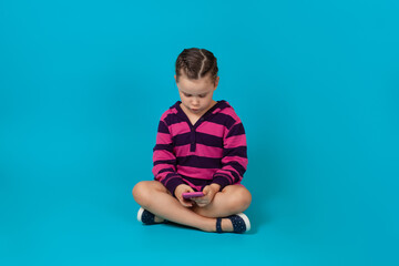 full-length portrait of an enthusiastic girl in a striped hoodie sitting cross-legged on the floor and playing games on a smartphone, isolated on a blue background.