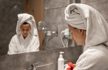 A young woman in a robe and with a towel on her head washes her hands in the sink.