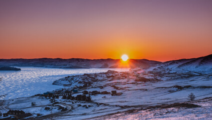 Winter sunrise. A sky of orange hues. The rays of the rising sun illuminate the blue frozen lake. On the snowy ground, between the hills, a small village is visible. Lake Baikal. Russia