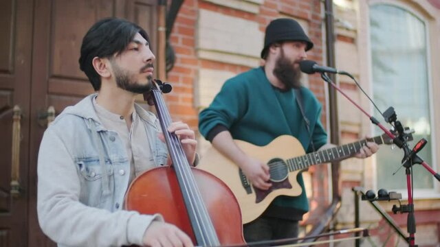 Medium shot of male musicians singing and playing acoustic guitar and cello on street