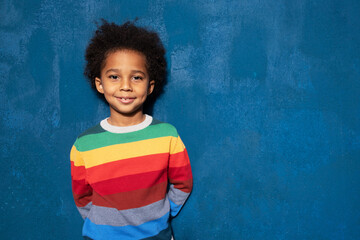 Cheerful african american little boy curly hairdo and dimples on cheeks wearing colorful sweater. Positive biracial schoolboy looking at camera with happy smile on blue background, copy space for ad.