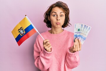 Young brunette woman holding colombia flag and colombian pesos banknotes puffing cheeks with funny face. mouth inflated with air, catching air.